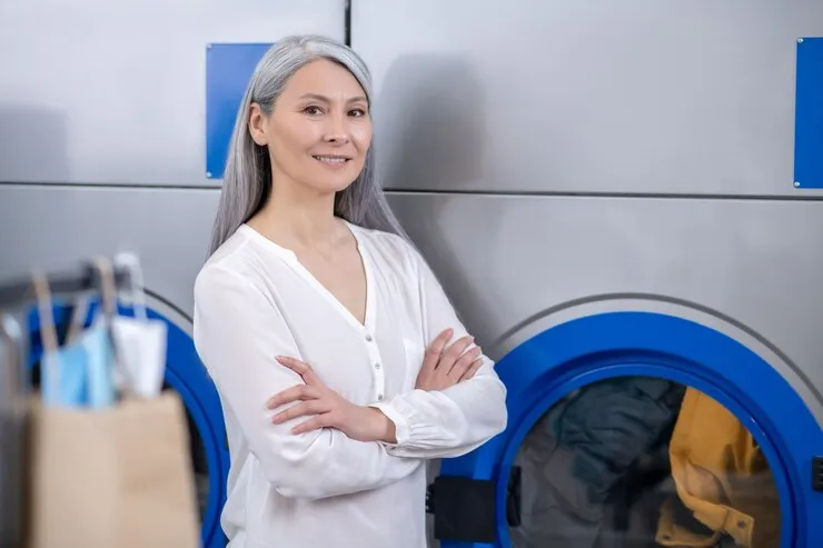 A woman with long gray hair and a white shirt stands with arms crossed in front of industrial washing machines at a laundromat.