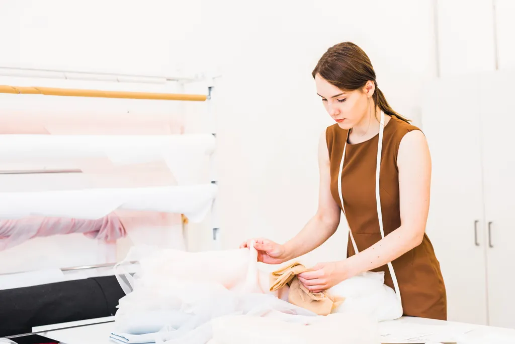 A woman in a brown dress measures and examines fabric samples on a table in a brightly lit workspace.
