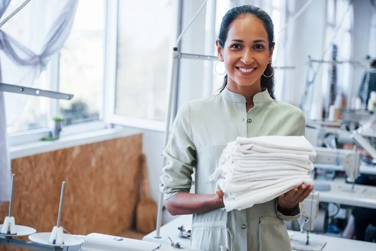Woman standing in a sewing workshop holding a folded stack of white fabric, smiling at the camera.
