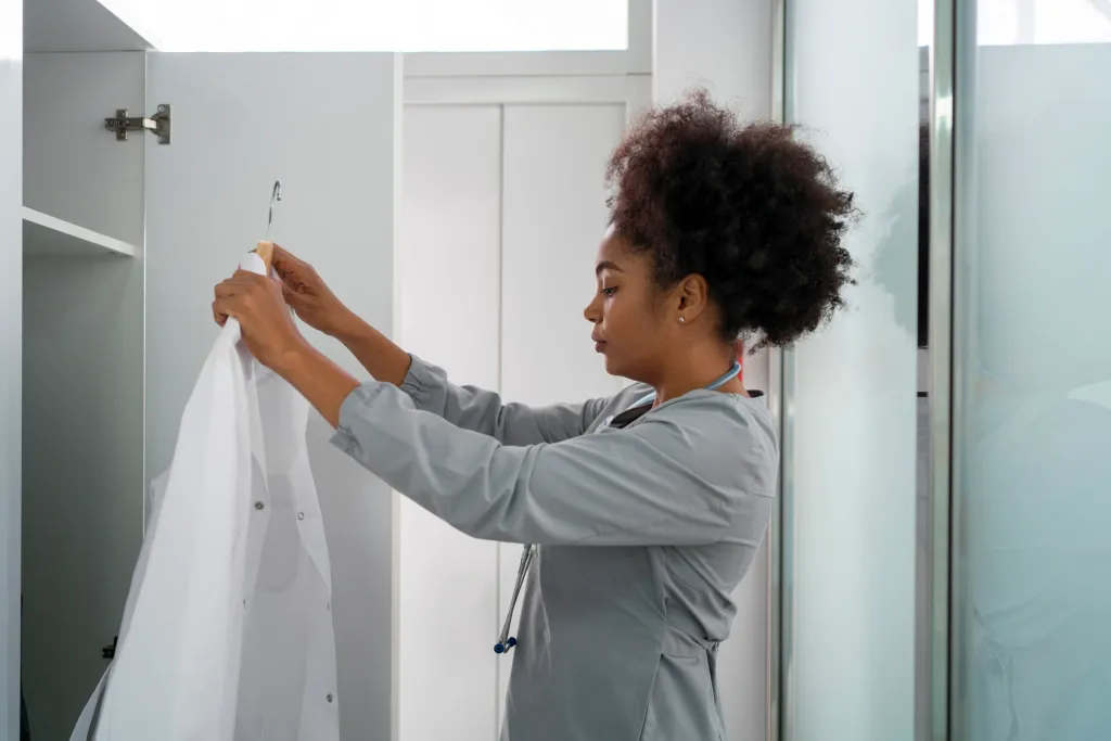 A woman wearing scrubs stands in front of an open locker, holding up a white lab coat on a hanger.