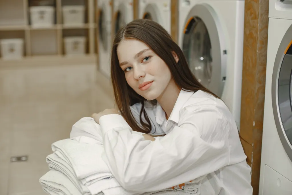 A woman in a white shirt sits in a laundromat, leaning on a stack of folded white towels in front of washing machines.