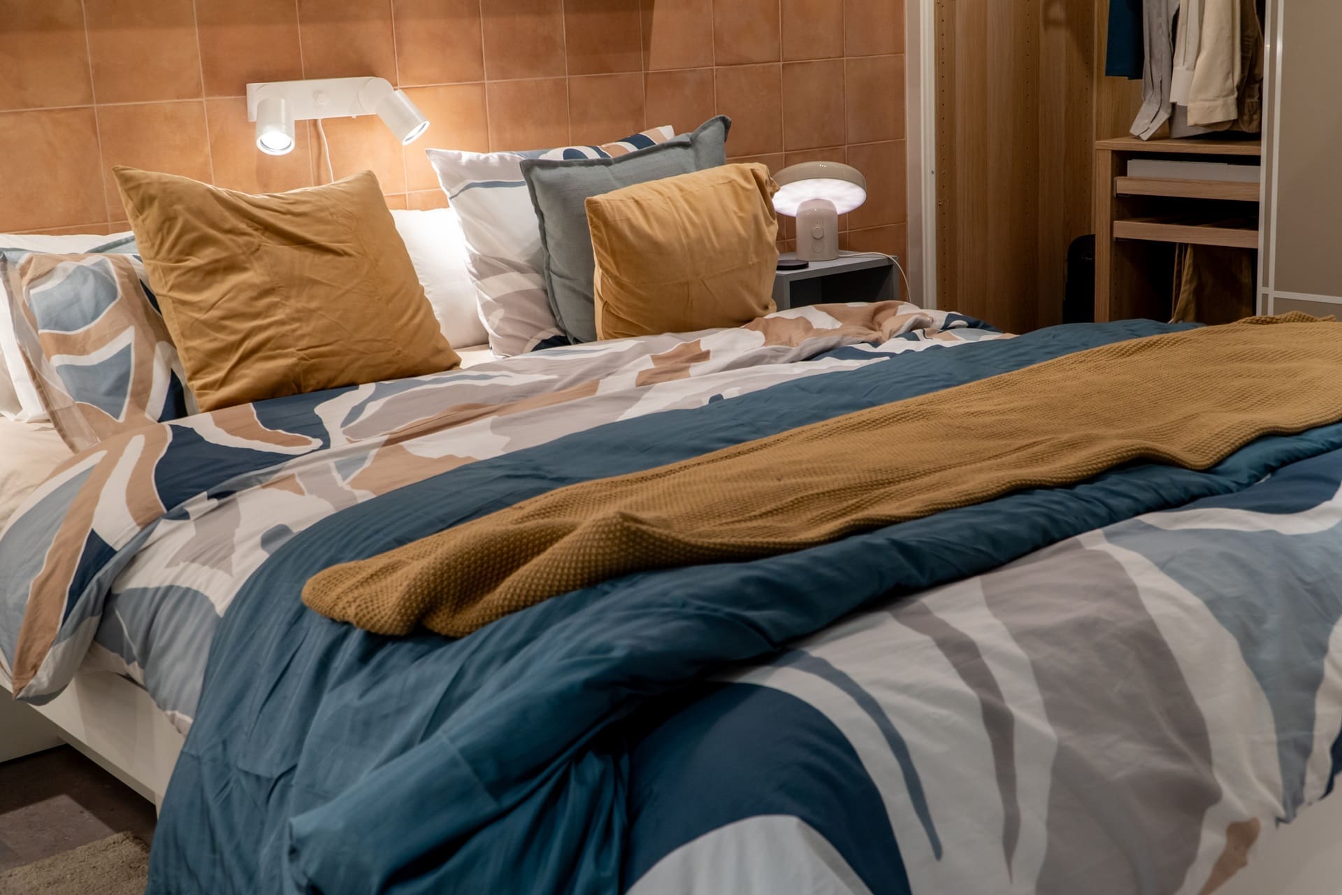 A neatly made bed with patterned blue, beige, and white bedding, mustard pillows and throw, a lamp on the nightstand, and a closet visible in the background.