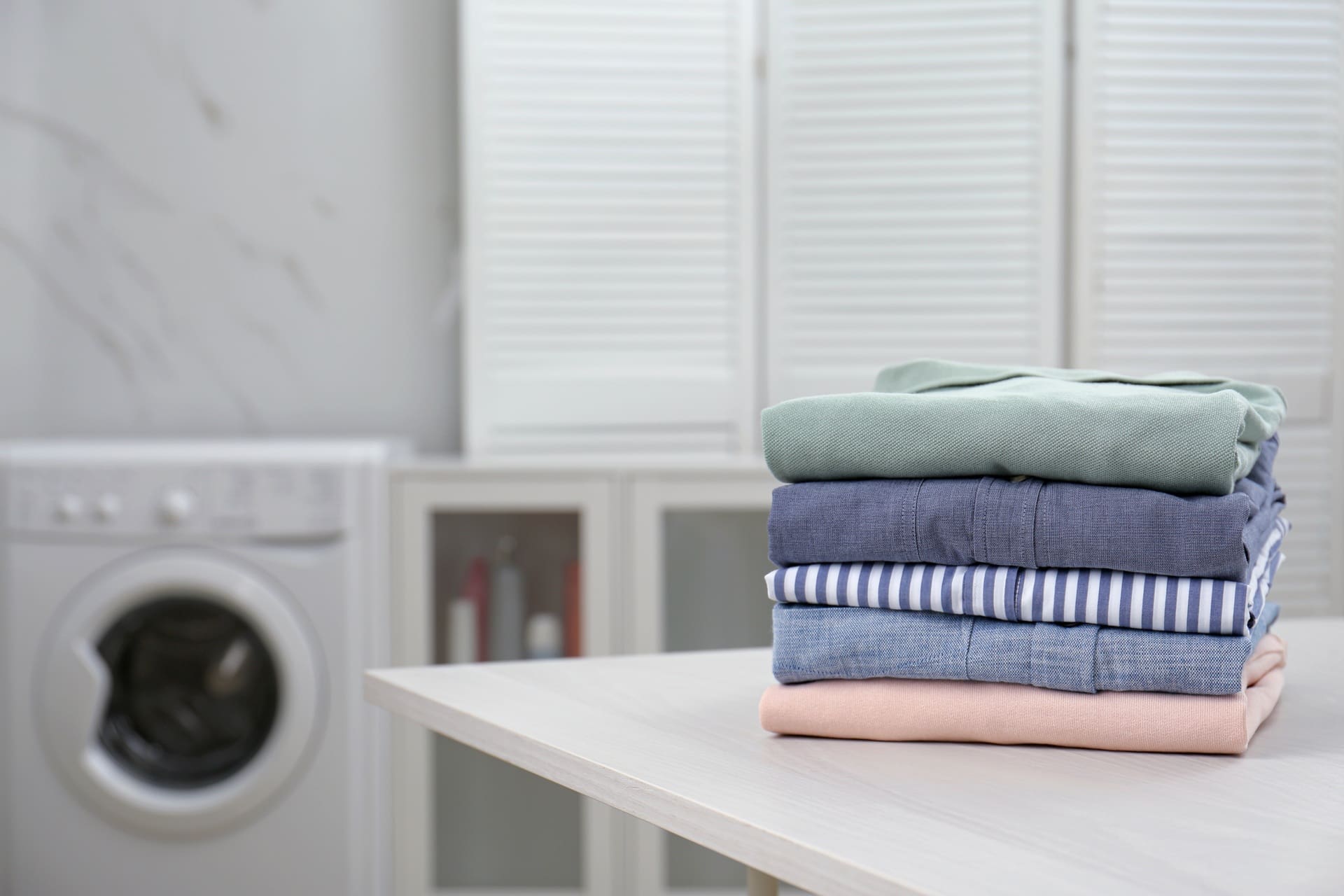 A neatly folded stack of clothes sits on a table, with a washing machine and cabinets visible in the background—showcasing the quality you can expect from a local laundry service Katy TX.