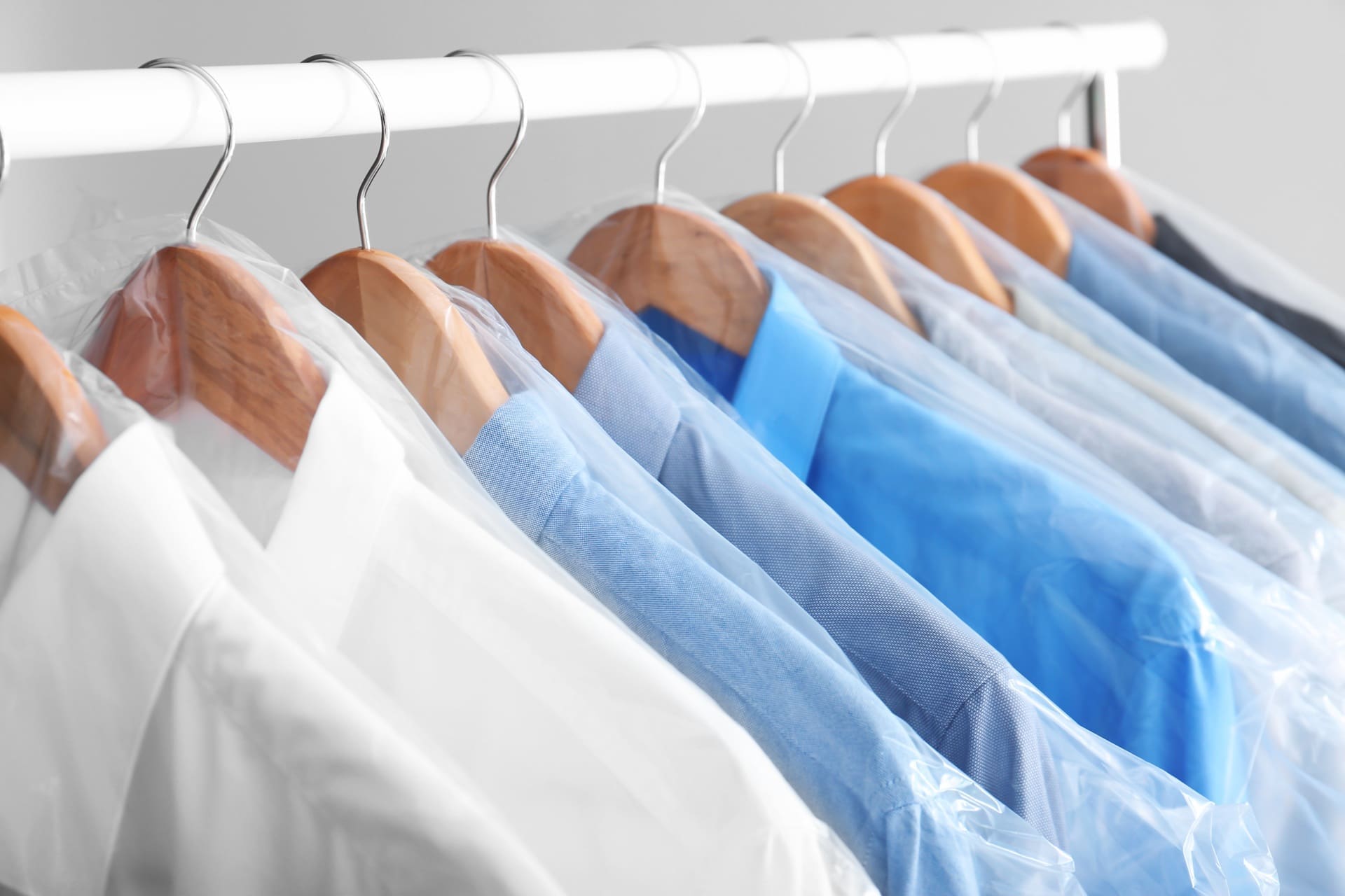 Several shirts in shades of blue and white hang on wooden hangers, each covered in plastic, on a white clothing rack—ready for pickup and delivery dry cleaning Katy.