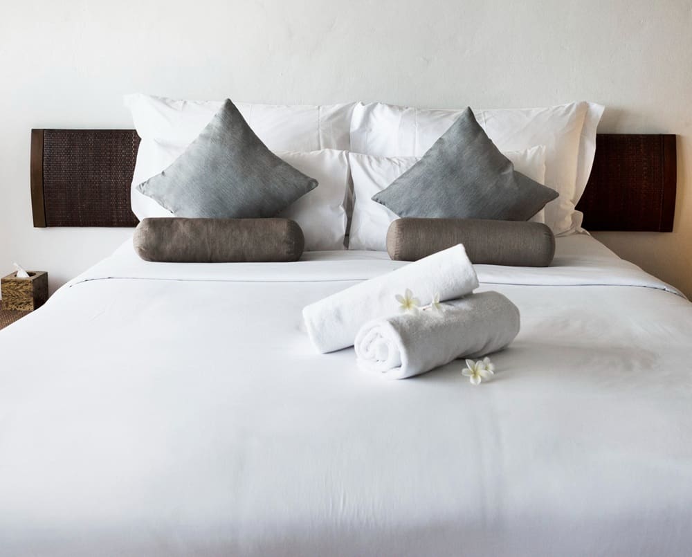 A neatly made bed with white sheets, two grey square pillows, two cylindrical brown cushions, and two rolled white towels with flowers on top—perfect for those searching for laundry near me or pickup and delivery laundry Katy.