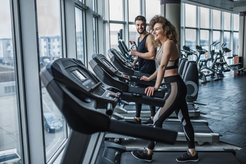 Two people in athletic wear use treadmills at a modern gym with large windows and various exercise equipment in the background.