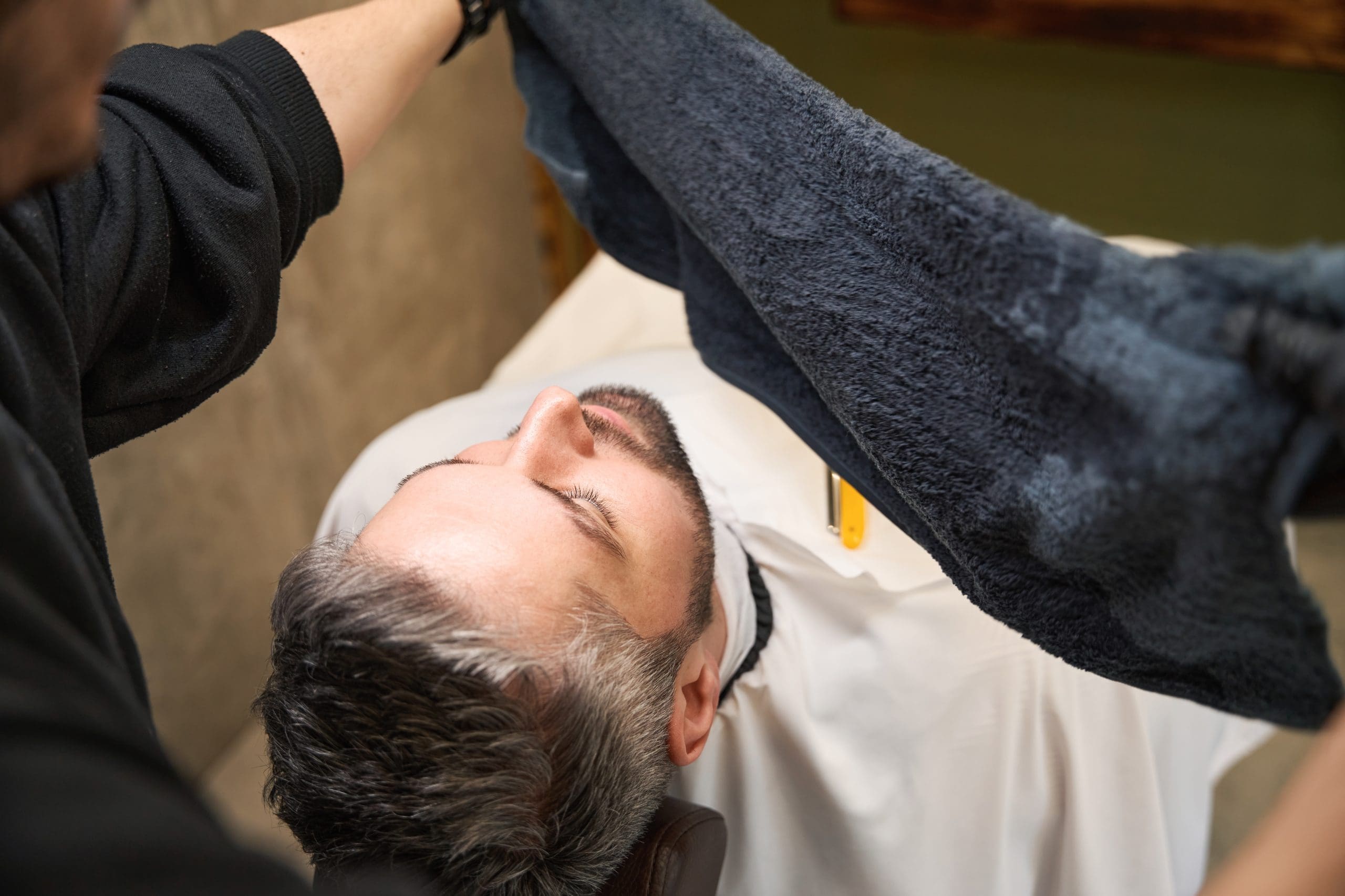 A man reclines in a barber chair with his eyes closed while a barber prepares to place a warm towel over his face.