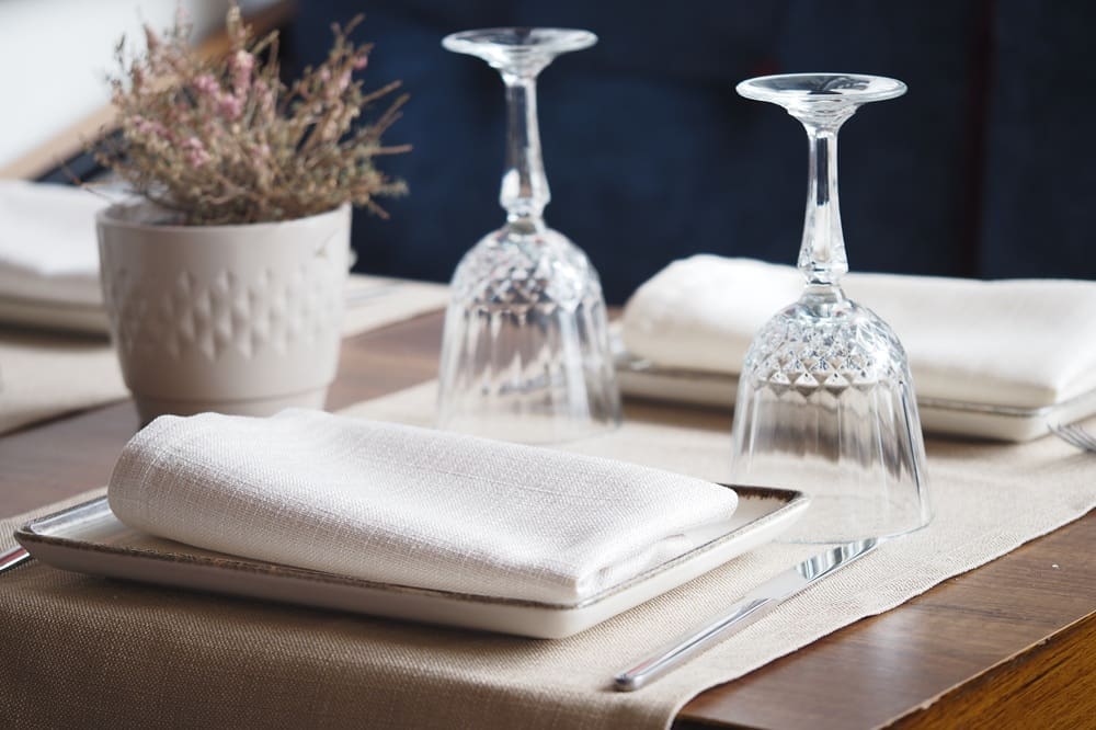 Table setting with two plates, each with a neatly folded white napkin, two upside-down wine glasses, cutlery, and a potted plant centerpiece on a wooden table.