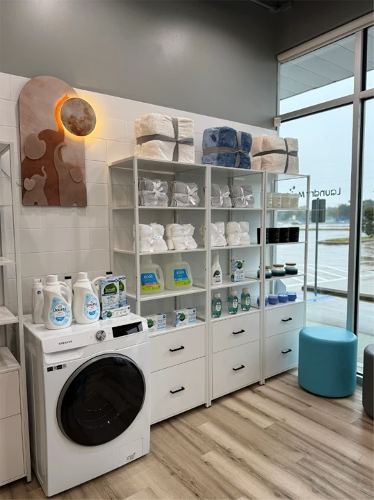 Shelves displaying laundry detergents, fabric softeners, and neatly folded towels next to a front-load washing machine in a modern, well-lit laundry facility.