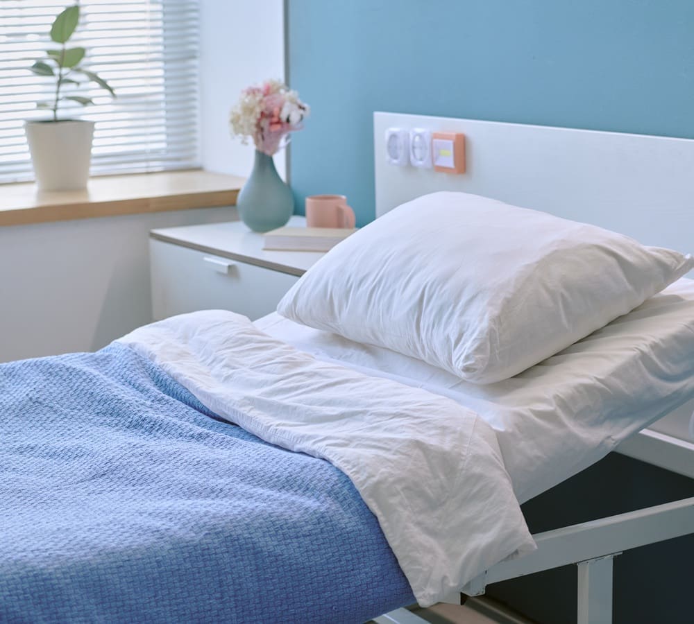 A neatly made hospital bed with white sheets and a blue blanket, beside a bedside table holding a vase of flowers, a plant, books, and a mug.
