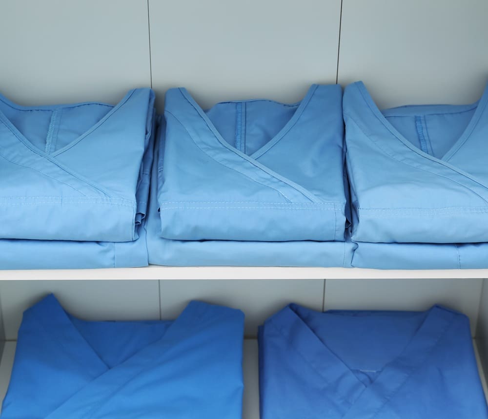 Neatly folded blue medical scrubs are arranged on white shelves, with three stacks on the top shelf and two on the bottom.