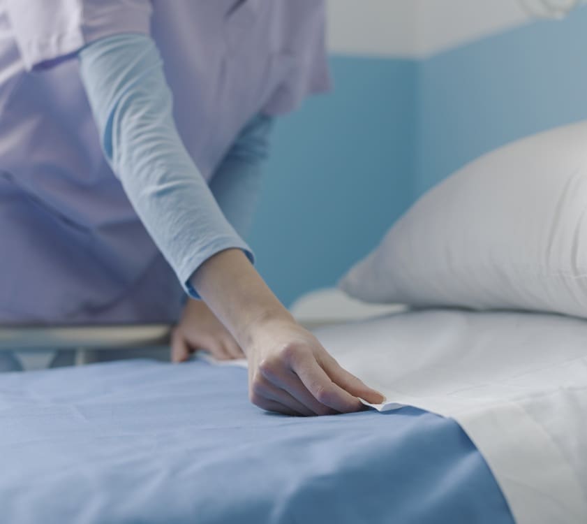 A person wearing scrubs makes a hospital bed, smoothing a blue sheet next to a white pillow in a clinical setting.