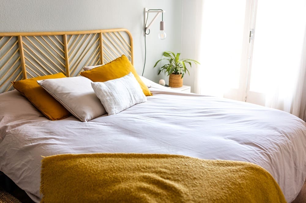 A neatly made bed with white linens, yellow cushions, and a mustard blanket, a rattan headboard, a wall-mounted lamp, and a potted plant on a bedside table near a window.