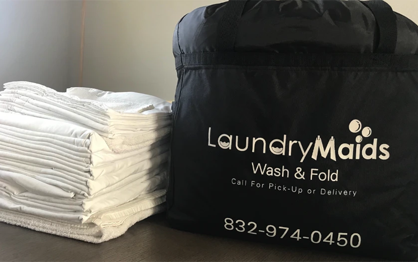 A black Laundry Maids bag sits next to a neatly folded stack of white towels on a wooden surface.